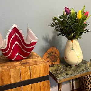 Può includere: Una ciotola di vetro a strisce rosse e bianche con un bordo ondulato si trova su un tavolo di legno. Un vaso con un motivo geometrico contiene un mazzo di tulipani viola, gialli e rosa.