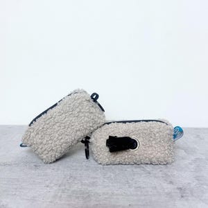 Könnte beinhalten: Zwei graue, flauschige, rechteckige Spender für Hundekotbeutel. Jeder hat einen schwarzen Reißverschluss, einen schwarzen Zuglasche und eine kleine schwarze Öffnung zur Ausgabe der Beutel. Einer hat eine blaue Zuglasche.