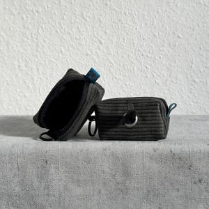 Könnte beinhalten: Zwei dunkelgraue Cord-Etuis mit gerippter Texturoberfläche. Ein Etui steht aufrecht und ist geöffnet, während das andere geschlossen horizontal liegt. Beide verfügen über ein kleines blaues Stoffetikett und eine schwarze Metallringbefestigung.