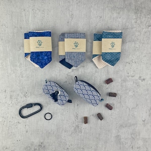 Könnte beinhalten: Drei blaue und weiße gemusterte Hundehalstücher mit brauner Papierverpackung, zwei blaue und weiße gemusterte Kotbeutelspender, ein schwarzer Karabinerhaken und ein schwarzer Ring sowie braune Hundekekse auf grauem Hintergrund.