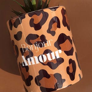 Peut inclure: Un pot de fleurs cylindrique avec un motif léopard dans les tons marron et noir. Les mots "Bonjour Amour" sont imprimés en blanc. Le pot contient une petite plante verte aux feuilles pointues.