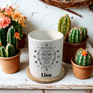 Peut inclure: Mug en céramique blanche avec un dessin de lion noir, entouré d'étoiles et de formes géométriques. Le mot "Lion" est imprimé en dessous. Le mug est posé sur un dessous de verre en bois, entouré de cactus et de fleurs en pot.