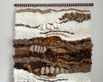 May include: A woven wall hanging in shades of brown, beige and white. The hanging features a variety of textures and patterns, including woven knots, fringe and burlap.