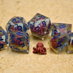 May include: A set of polyhedral dice made of clear resin, each containing a miniature red octopus figurine on a sandy base. The dice feature deep blue numbers. A small, loose red octopus figurine is positioned in the center of the group.