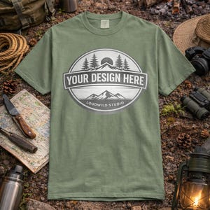 May include: An olive green t-shirt with a circular graphic featuring trees, mountains, and the text "YOUR DESIGN HERE" and "LOUDWILD STUDIO". The shirt is displayed on a natural outdoor setting with camping gear.