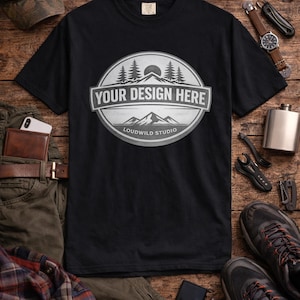 May include: Black t-shirt with a circular graphic that reads "YOUR DESIGN HERE" with a mountain and forest scene. The image includes a camouflage cap, sunglasses, watch, flask, shoes, and other accessories on a wooden surface.