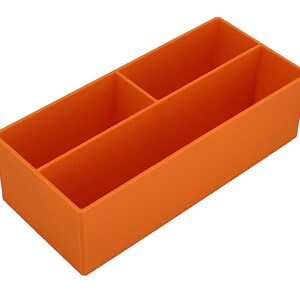 May include: An orange desk tidy with three compartments. The organiser is rectangular and made from a solid material. It is designed to hold pens, pencils, or other stationery items. The compartments are different sizes.