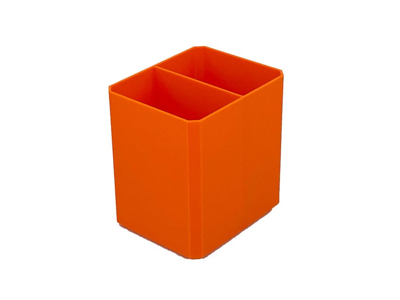 May include: Bright orange desk organizer with two compartments. The rectangular container is made of a solid material and is designed to hold pens, pencils, or other office supplies. The organizer is set against a white background.