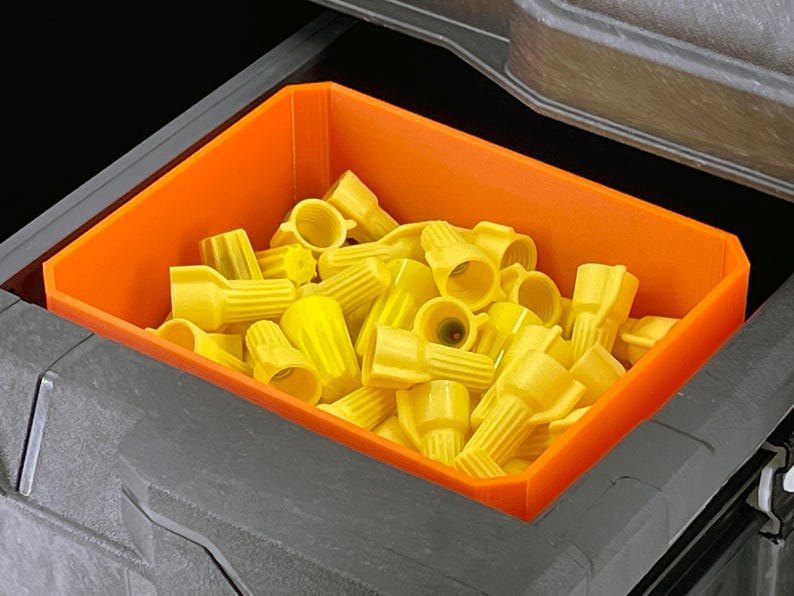 May include: An orange plastic bin filled with yellow wire connectors. The bin is rectangular and sits inside a larger gray container. The connectors are cone-shaped and have a ribbed texture.