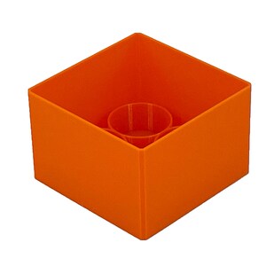 May include: An orange plastic cube with a round hole in the center. The cube is empty and appears to be a container.