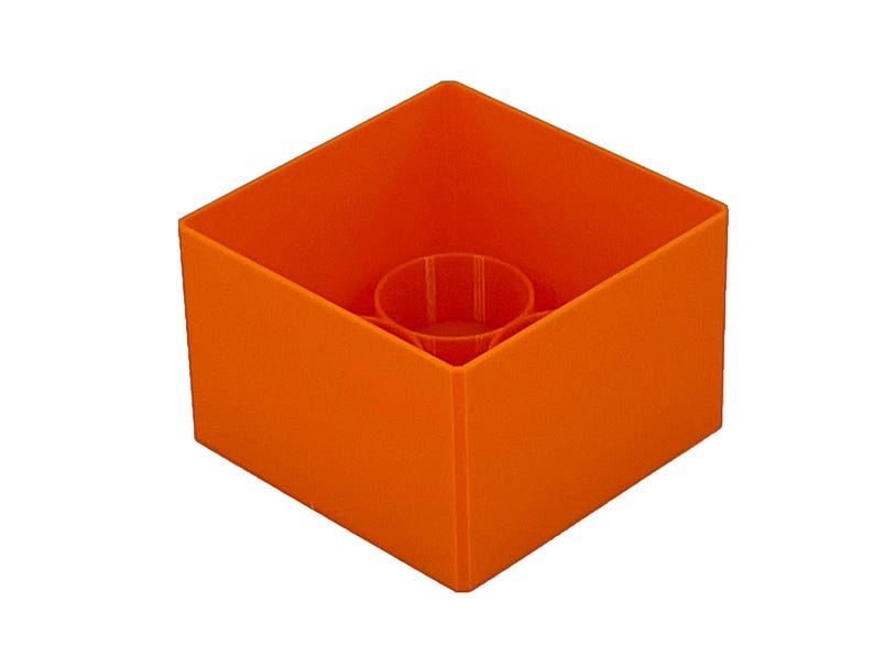 May include: An orange plastic cube with a round hole in the center. The cube is empty and appears to be a container.