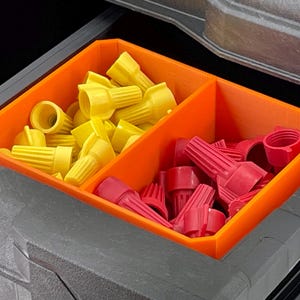 May include: An orange plastic organizer with two compartments, filled with yellow and red wire connectors. The organizer sits inside a dark gray storage container, providing a clear separation for electrical components.