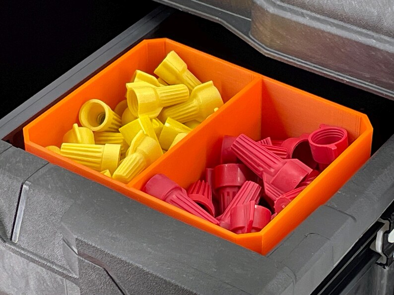 May include: An orange plastic organizer with two compartments, filled with yellow and red wire connectors. The organizer sits inside a dark gray storage container, providing a clear separation for electrical components.
