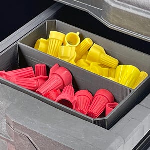 May include: A gray plastic organizer with two compartments, filled with red and yellow wire connectors. The connectors are various sizes and shapes, designed for electrical wiring projects. The organizer is inside a larger gray container.