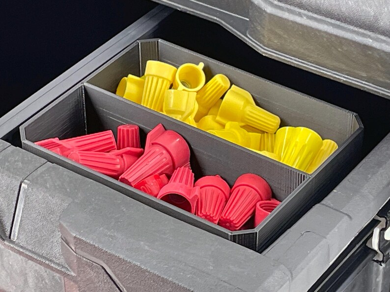 May include: A gray plastic organizer with two compartments, filled with red and yellow wire connectors. The connectors are various sizes and shapes, designed for electrical wiring projects. The organizer is inside a larger gray container.