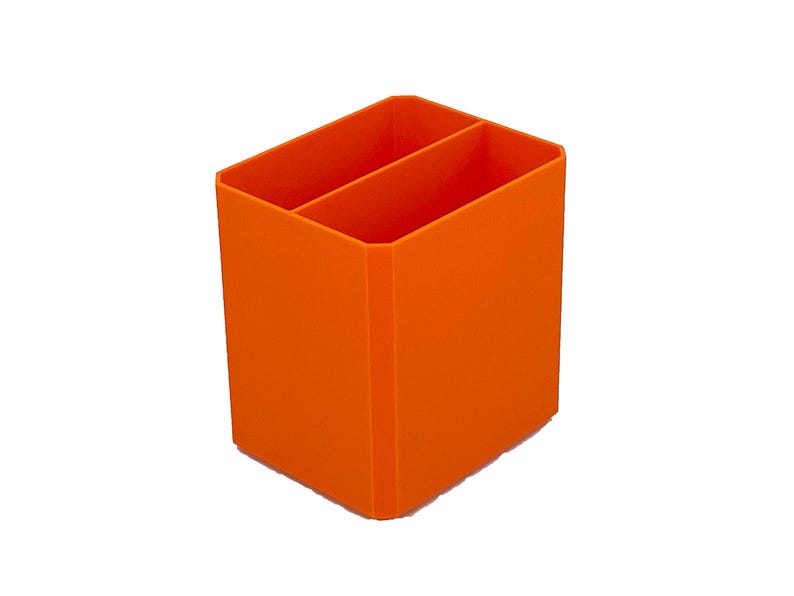 May include: Bright orange desk organizer with two compartments. The rectangular container is made of a solid material and is designed to hold pens, pencils, or other office supplies. The organizer is shown against a white background.