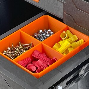 May include: An open drawer with an orange organizer containing various hardware items. The compartments hold screws, bolts, wire connectors, and other small parts. The drawer is part of a larger black storage system.