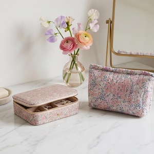 Könnte beinhalten: Eine rosa Schmuckschatulle und eine passende Kosmetiktasche mit Blumenmuster liegen auf einer Marmoroberfläche. Die Schmuckschatulle ist geöffnet und zeigt Fächer für Ringe und anderen Schmuck. Auf der Kosmetiktasche ist der Name "Sarah" gestickt. Eine Vase mit Blumen und ein goldener Spiegel sind im Hintergrund.