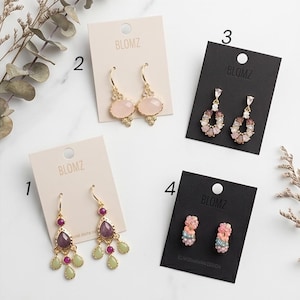Könnte beinhalten: Vier Paar Ohrringe auf Karten mit dem Markennamen "BLOMZ". Die Stile umfassen goldfarbene Creolen mit rosa Steinen, baumelnde Modelle mit lila und grünen Steinen sowie kleine florale Ohrstecker.