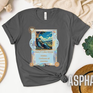 May include: A gray t-shirt with a graphic of a wizard lying down in a forest scene. The graphic is surrounded by a gold frame with a blue and pink background. The text on the graphic reads "Zero F*cks Given" and "Hidden. Confuses all enemies."