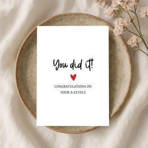 May include: A white rectangular greeting card with black text that reads You did it! and CONGRATULATIONS ON YOUR A-LEVELS, featuring a small red heart icon centred between the two lines of text on the front of the card.
