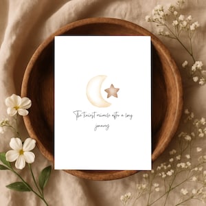 May include: A rectangular white card featuring a watercolour illustration of a pale yellow crescent moon and a light brown star. Below the celestial graphic, the black text reads: The Tiniest miracle after a long journey, written in a cursive script.