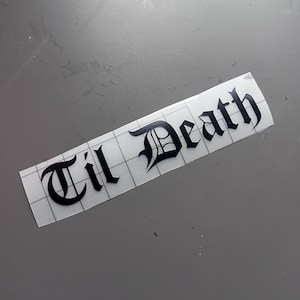 May include: A black vinyl sticker featuring the phrase "Til Death" in a gothic typeface. The sticker is rectangular, with a grid pattern on the backing, and is set against a grey background.