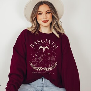 May include: Maroon sweatshirt with a graphic design featuring the text "BASGIATH WAR COLLEGE" above a mountain scene with a castle, stars, and a winged figure. The text "In the face of fear, we find our greatest courage" is below the graphic.