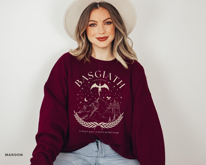 May include: Maroon sweatshirt with a graphic design featuring the text "BASGIATH WAR COLLEGE" above a mountain scene with a castle, stars, and a winged figure. The text "In the face of fear, we find our greatest courage" is below the graphic.