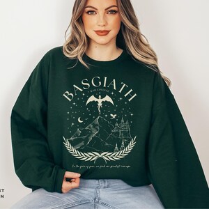 May include: Forest green sweatshirt with a graphic design featuring the word "BASGIATH", a winged creature, mountains, a castle, and the text "In the face of fear, we find our greatest courage."