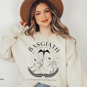 May include: Sand-colored sweatshirt with a black graphic featuring the text "BASGIATH WAR COLLEGE" above a mountain scene with a dragon, stars, and a castle. The text "In the face of fear, we find our greatest courage" is below the graphic.