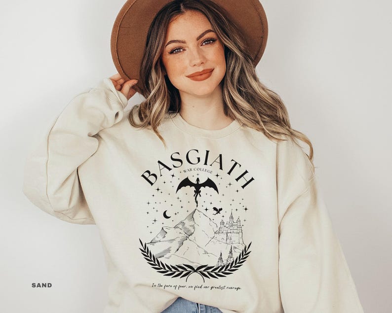 May include: Sand-colored sweatshirt with a black graphic featuring the text "BASGIATH WAR COLLEGE" above a mountain scene with a dragon, stars, and a castle. The text "In the face of fear, we find our greatest courage" is below the graphic.