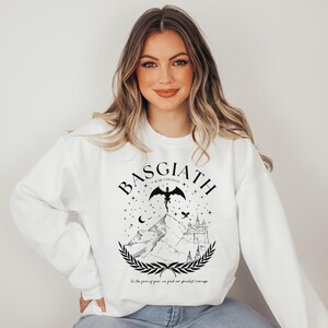 May include: White sweatshirt with a black graphic design featuring the word "BASGIATH" above a mountain scene with a dragon, stars, and a castle. The text "In the face of fear, we find our greatest courage" is below the graphic.