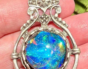 May include: A silver pendant with an iridescent blue gemstone. The pendant has an ornate, filigree design with floral accents. The gemstone is round and displays a vibrant array of colors, including blue, green, and red.