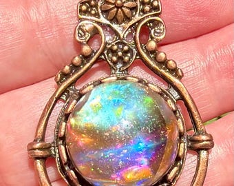 May include: Ornate bronze pendant featuring a large, iridescent, round stone with a spectrum of colors. The pendant has intricate filigree detailing, including a floral element at the top and decorative beading.