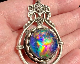 May include: A silver-toned pendant featuring an ornate, decorative metal frame with intricate scrollwork and floral details. The center holds a circular, domed cabochon displaying a vibrant, iridescent spectrum of blue, purple, and rainbow colors.