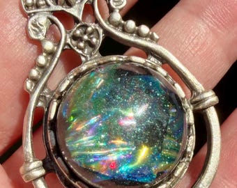 May include: A silver-toned metal pendant featuring an ornate, filigree-style top section with floral and beaded details. The center holds a circular, domed cabochon with a dark background and iridescent, multi-colored flecks of green, blue, and gold.