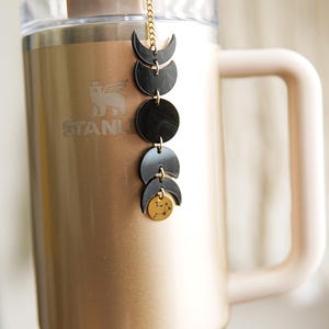 May include: A metallic gold tumbler with a handle and a clear straw, featuring a hanging charm accessory. The charm consists of a gold chain with black acrylic moon phase shapes and a small circular gold pendant with a constellation design.