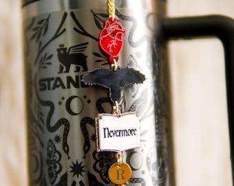 May include: A metallic gray tumbler with a black handle and a dark graphic pattern featuring skulls, snakes, and moths. A gold chain charm hangs from the lid, displaying a red anatomical heart, a black raven, an open book labeled Nevermore, and a gold disc with the letter R.
