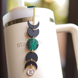 May include: A gold chain with black and green moon and circle charms hangs from a white travel mug with the word "Stanley" printed on it. The charm at the bottom of the chain has the letter "E" on it.