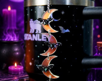 May include: A black tumbler featuring a silver chain accessory with three orange crescent moon charms, each adorned with a small black glittery bat. A circular charm with the letter K is attached to the bottom of the chain. The tumbler has the Stanley logo printed in white on the side.