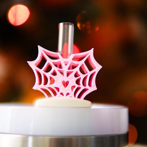 May include: A light pink, 3D-printed spider web straw topper featuring a small heart shape at its center. The topper is attached to a clear plastic straw, which is inserted into the white lid of a tumbler. The design is symmetrical and intricate.