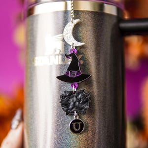 May include: A silver and black travel mug with a decorative charm. The charm features a crescent moon, a witch's hat, a cauldron, and a charm with the letter "U". The mug has the word "STANLEY" printed on it.