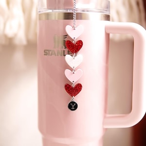 Könnte beinhalten: Ein rosa isolierter Becher mit Henkel und transparentem Deckel. Ein dekorativer Anhänger mit roten, weißen und rosa herzförmigen Anhängern hängt am Deckel. Der Anhänger endet mit einem schwarzen runden Anhänger mit einem weißen "Y".