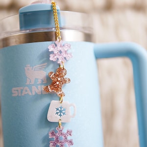 Puede incluir: Una botella de agua de acero inoxidable azul claro con una cadena dorada y abalorios. Los abalorios incluyen un hombre de jengibre, un copo de nieve y una taza de caf&eacute;. La botella tiene la palabra "STANLEY" impresa en el lateral.