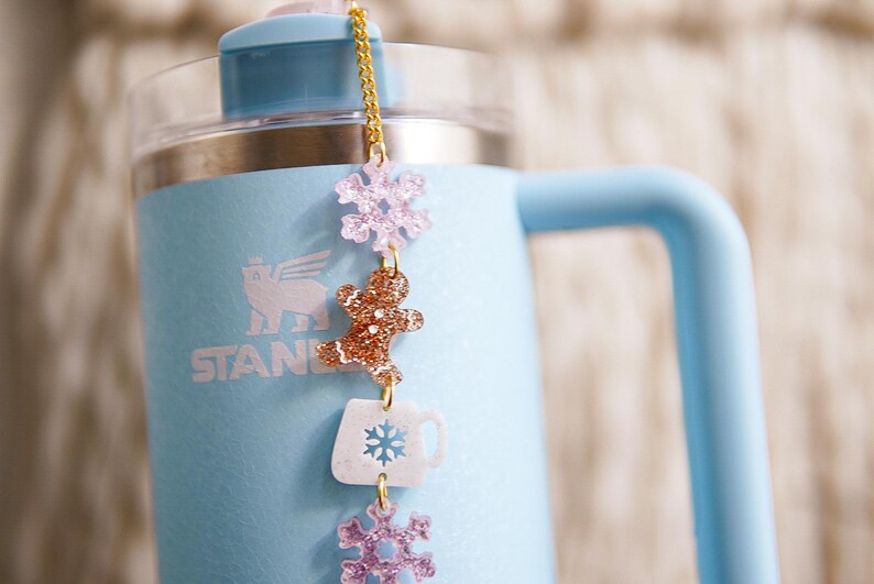 Puede incluir: Una botella de agua de acero inoxidable azul claro con una cadena dorada y abalorios. Los abalorios incluyen un hombre de jengibre, un copo de nieve y una taza de caf&eacute;. La botella tiene la palabra "STANLEY" impresa en el lateral.