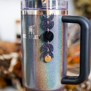 May include: A metallic gray Stanley tumbler with a black handle and a clear straw, featuring a vertical charm chain attached to the lid. The charm displays a sequence of four moon phases in dark glittery purple and black, ending with a small gold disc.
