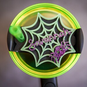 May include: A green and black spiderweb-shaped lid with the name "Savannah" written in purple glitter. The lid is on top of a green and black tumbler.