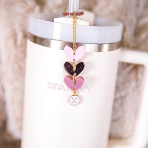 May include: White stainless steel travel mug with a black and pink heart charm hanging from the lid. The charm is attached to a gold chain and has a small white circle with an 'X' on it.