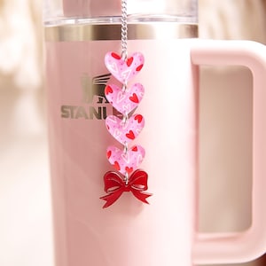Könnte beinhalten: Ein rosa isolierter Becher mit Griff und silbernem Logo. Ein dekorativer Anhänger hängt am Deckel, mit rosa Herzen mit roten Herzmustern und einer roten Schleife. Der Becher ist für Getränke konzipiert.